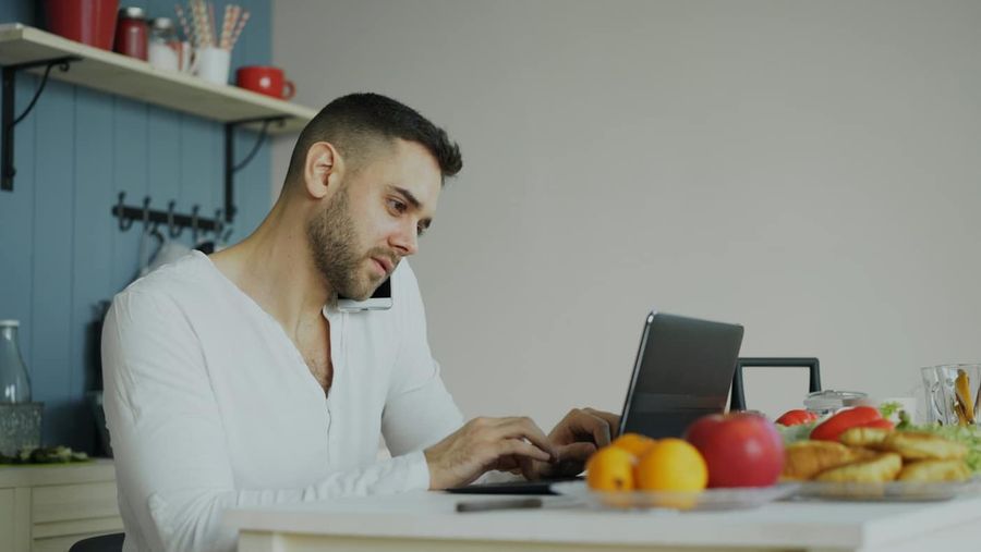 Mężczyzna rozmawia przez telefon, pracując na laptopie w kuchni
