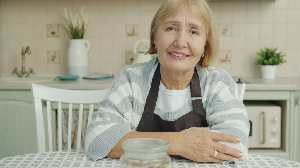 Starsza kobieta siedzi przy kuchennym stole, w tle czajnik i kuchenka